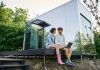 young multiethnic couple sitting on porch with laptop near modern glass house in countryside