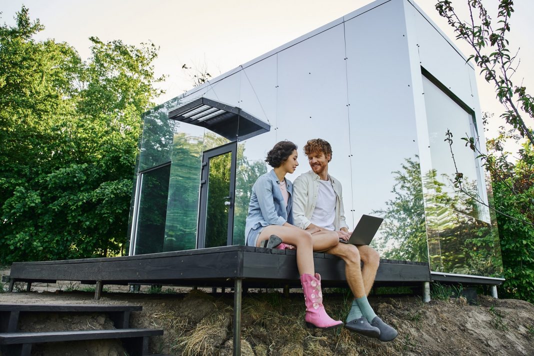 young multiethnic couple sitting on porch with laptop near modern glass house in countryside