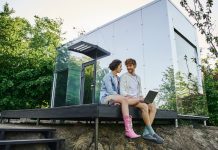 young multiethnic couple sitting on porch with laptop near modern glass house in countryside