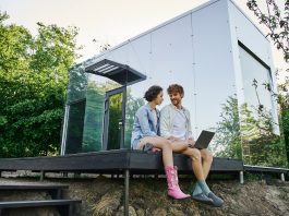 young multiethnic couple sitting on porch with laptop near modern glass house in countryside