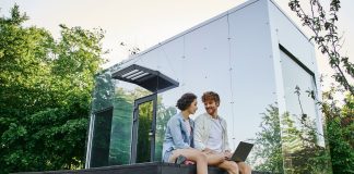 young multiethnic couple sitting on porch with laptop near modern glass house in countryside