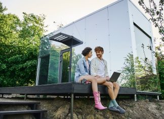 young multiethnic couple sitting on porch with laptop near modern glass house in countryside