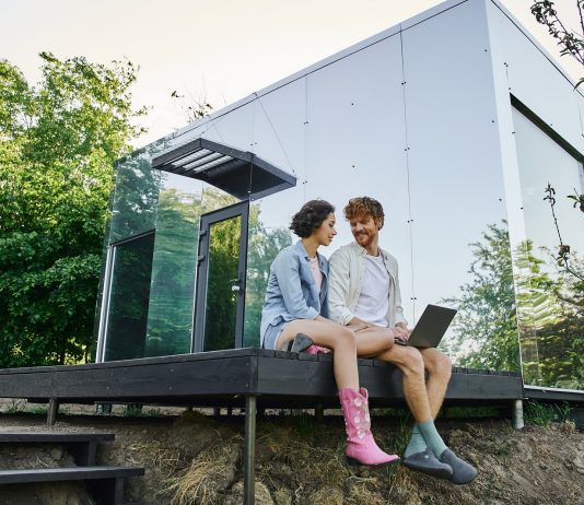 young multiethnic couple sitting on porch with laptop near modern glass house in countryside
